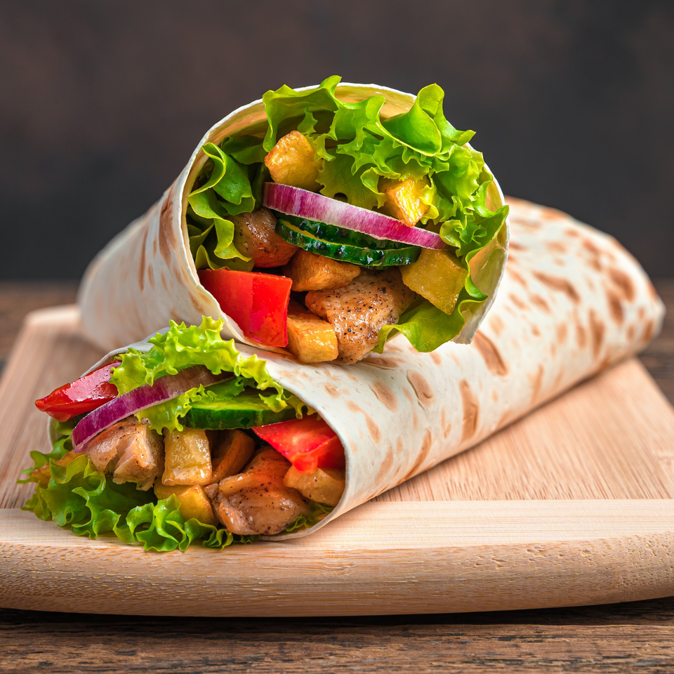 Two chicken wraps filled with lettuce, red onion, cucumber, tomatoes, and golden potatoes on a wooden board.