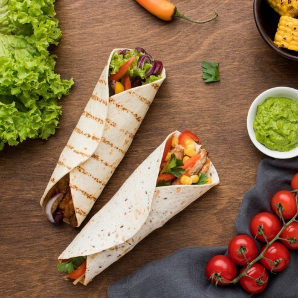 Two grilled chicken wraps with fresh vegetables, guacamole, and cherry tomatoes on a wooden table.