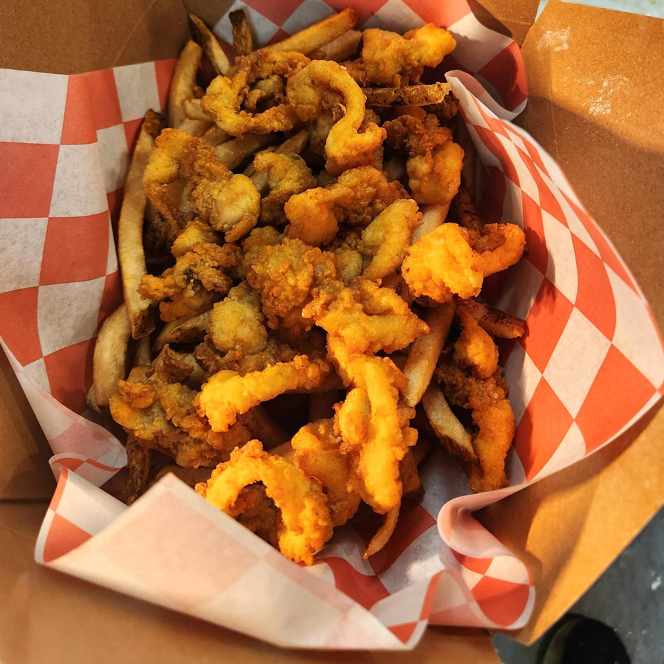 A cardboard container filled with golden-brown fried clams and french fries, lined with red and white checkered paper.